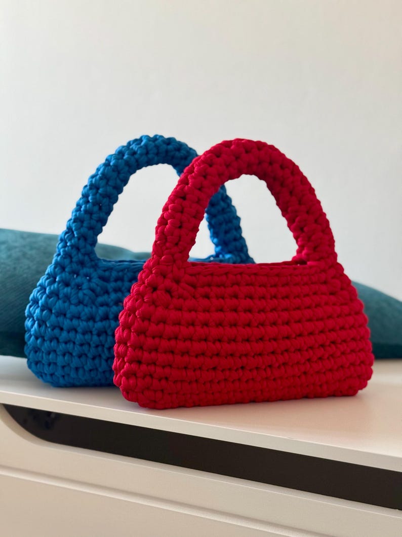 May include: Two crocheted handbags, one red and one blue. The bags have a simple, rectangular shape with a rounded top and a single handle.