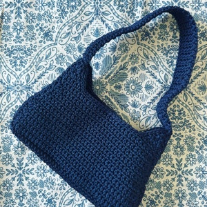 May include: A dark blue crocheted handbag with a single strap. The bag is on a white and blue patterned fabric.