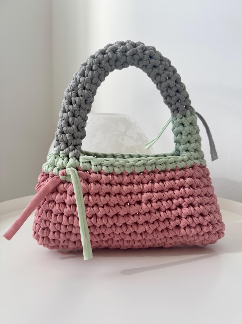 May include: A crocheted handbag with a gray, green, and pink color scheme. The bag has a single handle and a rectangular shape.