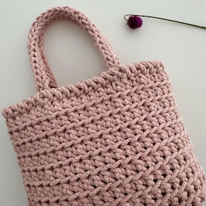 May include: A pink crocheted tote bag with a woven pattern. The bag has two handles and is made of a soft, textured yarn.