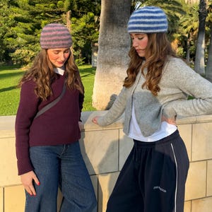 Op de afbeelding: Twee mensen met gestreepte gebreide mutsen. De ene muts is roze en grijs, de andere blauw en wit. De ene persoon draagt een bordeauxrode trui en een spijkerbroek, de andere een grijze cardigan en een zwarte joggingbroek.