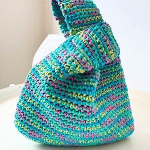 Op de afbeelding: Een handgehaakte tas in tinten van turquoise, roze en geel. De tas heeft een unieke geknoopte handgreep en een afgeronde bodem. De getextureerde steek creëert een visueel aantrekkelijk oppervlak. Deze kleurrijke tas is perfect voor het dragen van benodigdheden. De tas is ongeveer 25 cm hoog.