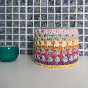 May include: A handmade, colourful crochet toilet paper cover. The cover features a rainbow of pastel colours, including pink, yellow, orange, blue, and purple. The crochet pattern creates a textured, geometric design. A teal glass candle holder is to the left.