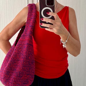 May include: A handmade, crochet shoulder bag in shades of red and purple. The bag has a rounded shape and a wide strap. The bag is made of a textured yarn and appears to be sturdy. The bag is being held over the shoulder.