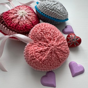 May include: Crocheted heart-shaped items in shades of pink, red, and gray, with satin ribbon loops. Accompanying the hearts are purple heart-shaped soaps and a red heart-shaped stone with a sun design.