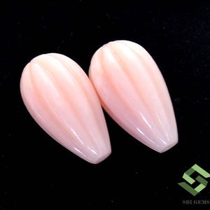 May include: Two light pink, carved gemstone beads. Each bead is elongated with a rounded top and a pointed bottom. The surface of each bead has vertical grooves. The beads are displayed against a black background.