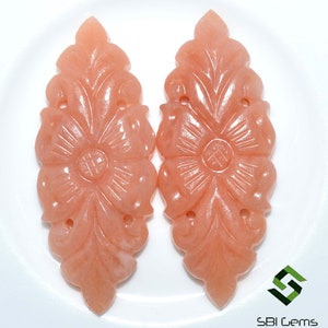 May include: Two peach-colored, carved gemstone pendants. Each pendant features a floral design with detailed leaves and a central flower. The pendants are symmetrical and have small holes for stringing. The background is white.
