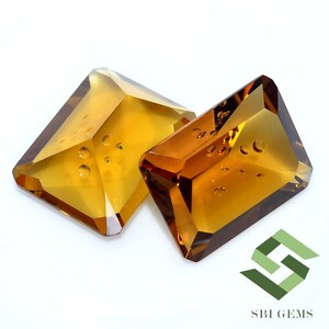 May include: Two faceted, rectangular-cut gemstones with a warm, amber hue. The gems have a geometric shape with multiple facets, catching the light. The gems are set against a white background. The logo "SBI GEMS" is in the bottom right corner.