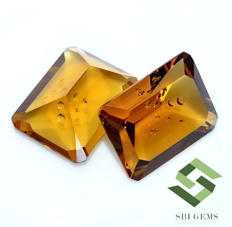 May include: Two faceted, rectangular-cut gemstones with a warm, amber hue. The gems have a geometric shape with multiple facets, catching the light. The gems are set against a white background. The logo "SBI GEMS" is in the bottom right corner.