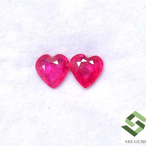 Könnte beinhalten: Zwei herzförmige, facettierte, leuchtend rosa Edelsteine. Die Edelsteine haben eine polierte Oberfläche und eine weiße, dreieckige Facette oben. Der Hintergrund ist ein weicher, weißer Stoff. Das Logo "SBI GEMS" befindet sich unten rechts.