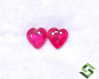 May include: Two heart-shaped, faceted, bright pink gemstones. The gems have a polished surface and a white, triangular facet at the top. The background is a soft, white fabric. The logo "SBI GEMS" is in the bottom right corner.