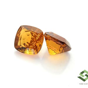 May include: Two faceted, golden-yellow gemstones on a white background. The larger gem has a cushion cut, while the smaller one is rectangular. The gems reflect light, showcasing their clarity and brilliance. The image is well-lit, highlighting the gems' color and facets.