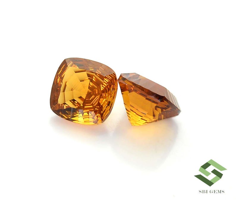 May include: Two faceted, golden-yellow gemstones on a white background. The larger gem has a cushion cut, while the smaller one is rectangular. The gems reflect light, showcasing their clarity and brilliance. The image is well-lit, highlighting the gems' color and facets.
