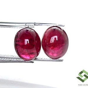 Könnte beinhalten: Zwei ovale, polierte Rubin-Edelsteine. Die Edelsteine haben eine tiefe, satte rote Farbe mit einer glatten, reflektierenden Oberfläche. Sie werden von einer Pinzette vor einem weißen Hintergrund gehalten. Die Edelsteine sind wahrscheinlich für die Schmuckherstellung.