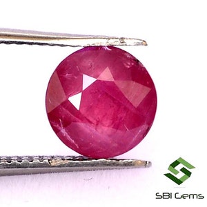 May include: A round, faceted ruby gemstone with a deep pink hue. The ruby is held in place by a pair of tweezers.