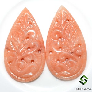 May include: Two peach-colored, teardrop-shaped gemstones with intricate carved floral and swirl designs. Each stone features a series of small, evenly spaced holes. The gemstones are displayed against a white background.
