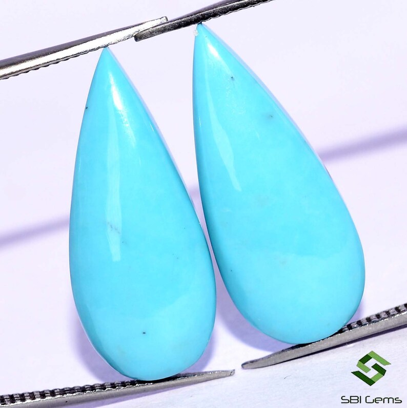 May include: Two teardrop-shaped turquoise gemstones, each with a smooth, polished surface and a vibrant blue color. The stones are held by tweezers against a white background.