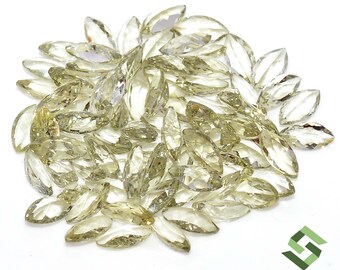 May include: A collection of marquise-cut gemstones in various shades of yellow and clear. The gemstones are scattered on a white surface, with light reflecting off their facets. The image is well-lit, showcasing the clarity and color of the stones.