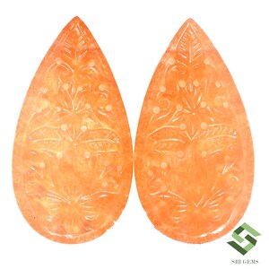 May include: Two orange-colored, teardrop-shaped gemstones with intricate floral carvings. The gemstones have a translucent quality, and the carvings feature leaves and small circular details. The background is white.