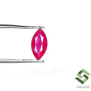 May include: A marquise-cut ruby gemstone with a vibrant red color and faceted surface, held by silver-toned metal tweezers. The gemstone displays a symmetrical elongated shape with pointed ends and internal light reflections.