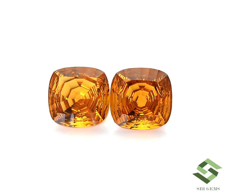 May include: Two cushion-cut, golden-yellow gemstones are displayed against a white background. The gems have a faceted surface that reflects light, creating a brilliant sparkle. The gems are a vibrant, warm color.