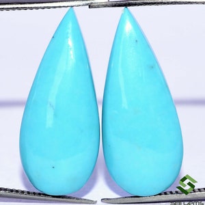 May include: Two teardrop-shaped turquoise cabochons, each measuring approximately 1 inch in length, are held by a pair of tweezers. The stones are a vibrant blue color with a smooth, polished surface.