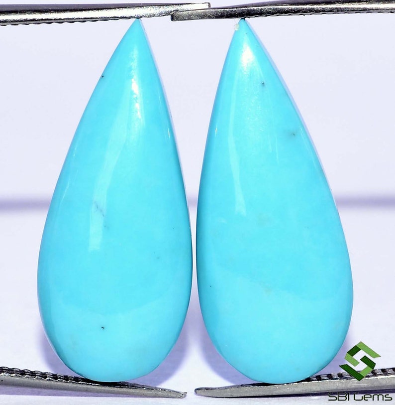 May include: Two teardrop-shaped turquoise cabochons, each measuring approximately 1 inch in length, are held by a pair of tweezers. The stones are a vibrant blue color with a smooth, polished surface.