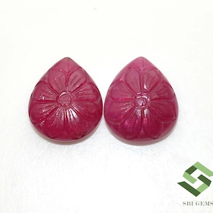 Puede incluir: Dos gemas talladas en forma de lágrima, de color magenta intenso. Cada gema presenta un diseño floral detallado tallado en la superficie. Las gemas se muestran sobre un fondo blanco.