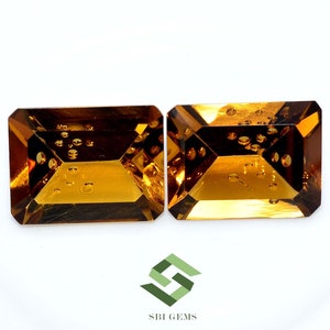 May include: Two rectangular, faceted gemstones with a warm, amber hue. The gems have a step-cut design and internal inclusions, set against a white background. The logo "SBI GEMS" is visible in the bottom right corner.