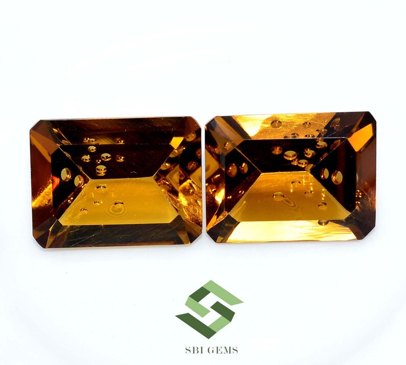 May include: Two rectangular, faceted gemstones with a warm, amber hue. The gems have a step-cut design and internal inclusions, set against a white background. The logo "SBI GEMS" is visible in the bottom right corner.