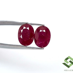 May include: Two oval-cut, deep red rubies are held by tweezers against a white background. The gemstones have a faceted surface, reflecting light. The image is well-lit, highlighting the colour and clarity of the rubies. The gems are a rich, saturated red.