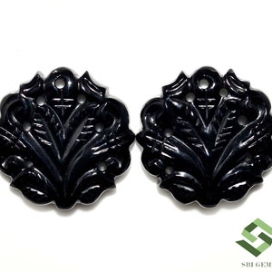 Könnte beinhalten: Zwei schwarze Onyx-Cabochons mit aufwendigen floralen Schnitzereien und mehreren Löchern. Die Cabochons sind rund und haben eine polierte Oberfläche.