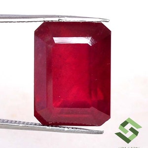 May include: A deep red gemstone with a rectangular emerald cut. The gemstone is held in tweezers.