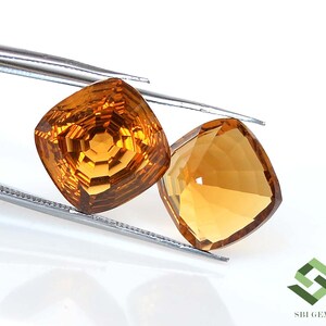 May include: Two faceted, cushion-cut gemstones in shades of amber and gold are held by a pair of tweezers. The gems have a brilliant, reflective surface, showcasing intricate internal facets. The background is white, highlighting the gems' color and clarity.