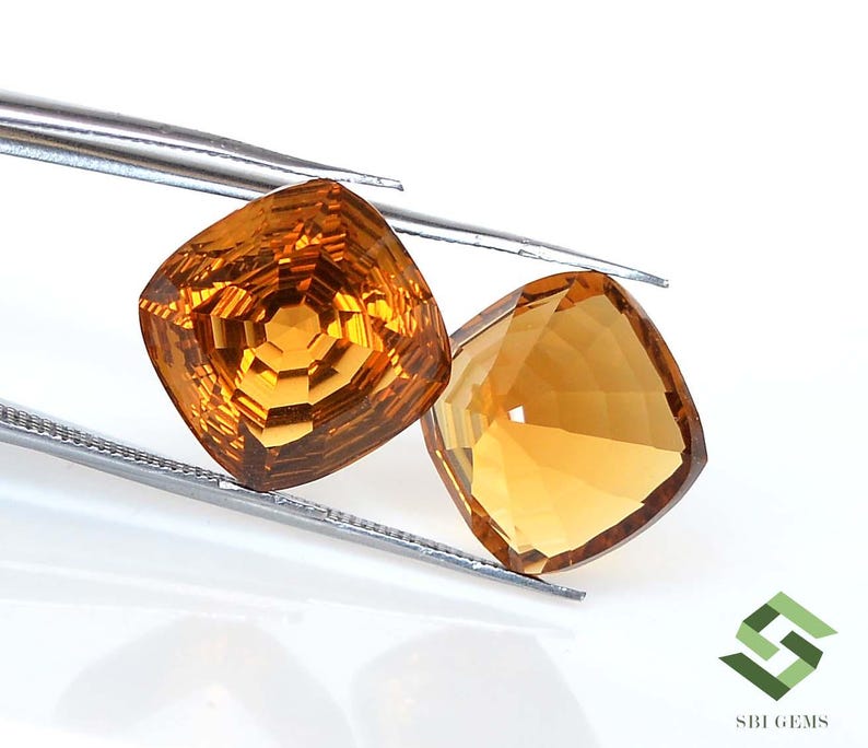 May include: Two faceted, cushion-cut gemstones in shades of amber and gold are held by a pair of tweezers. The gems have a brilliant, reflective surface, showcasing intricate internal facets. The background is white, highlighting the gems' color and clarity.