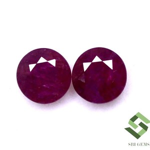 May include: Two round, faceted ruby gemstones with a deep red colour. The gemstones have a white, triangular marking on the top.