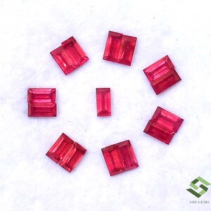May include: Eight red rectangular gemstones, also known as rubies, are arranged in a circular pattern on a white surface. The gemstones are all the same size and shape, and they are all cut in a rectangular shape.