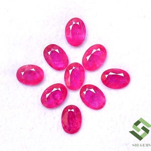 May include: Eight oval-cut, vibrant pink-red gemstones arranged on a white surface. Each gem has a faceted top, catching the light. The gems are likely rubies, showcasing a rich, saturated colour. The image includes the SBI GEMS logo.