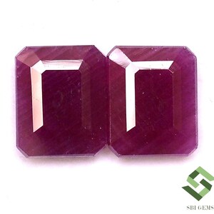 May include: Two emerald cut ruby gemstones, each with a deep red hue and a polished, faceted surface. The stones are set side-by-side, showcasing their symmetrical shape and brilliance.
