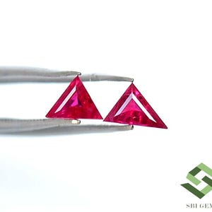 Puede incluir: Dos gemas triangulares, facetadas, de color rojo brillante, sujetas por unas pinzas plateadas sobre un fondo blanco. Las gemas tienen una forma geométrica afilada y una superficie pulida que refleja la luz.