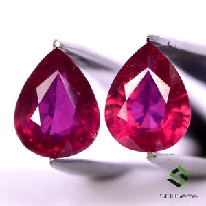 May include: Two pear-shaped ruby gemstones, each with a deep red color and a faceted cut. The gemstones are held by tweezers against a white background.