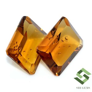 May include: Two faceted, amber-colored gemstones with a rectangular shape and beveled edges. The gems have internal inclusions and are set against a white background. The gems are likely for jewelry making.