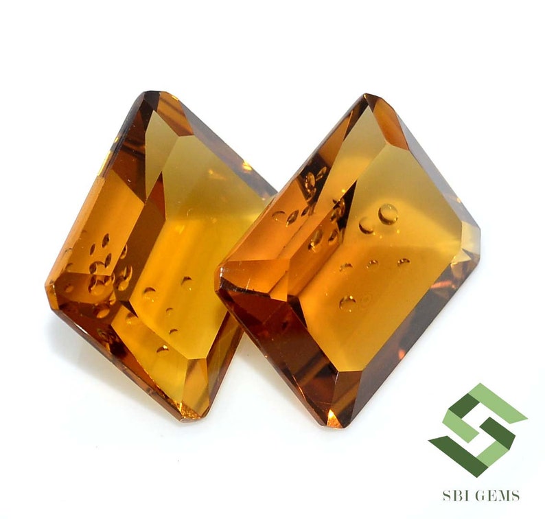 May include: Two faceted, amber-colored gemstones with a rectangular shape and beveled edges. The gems have internal inclusions and are set against a white background. The gems are likely for jewelry making.