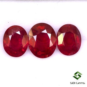 May include: Three oval-shaped red gemstones, likely rubies, are arranged in a row on a white surface. The gemstones are polished and have a glossy finish.