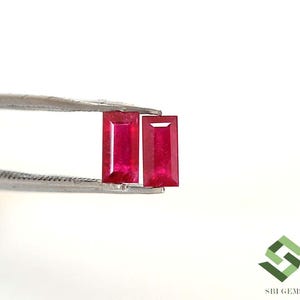 May include: Two vibrant red ruby gemstones, rectangular in shape, are held by a pair of tweezers against a white background. The gems are a deep, rich red color, showcasing their clarity and brilliance. The image highlights the gemstones' cut and color.
