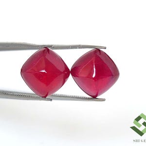 Puede incluir: Dos piedras preciosas de color rojo intenso, talla cojín, con una superficie lisa y pulida. Las piedras están sujetas por unas pinzas de metal plateado. En la esquina inferior derecha hay un logotipo geométrico verde con el texto SBI GEMS.