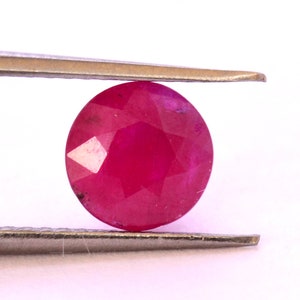 May include: A round, faceted ruby gemstone with a deep red color. The gemstone is held in place by a pair of tweezers.