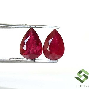 May include: Two faceted, pear-shaped rubies, held by tweezers, against a white background. The rubies are a deep, rich red colour, with visible facets that catch the light. The gems are likely for jewellery making.