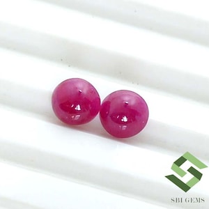 May include: Two round, polished, deep pinkish-red gemstone cabochons with a smooth, glossy surface. The stones are positioned side-by-side, displaying a vibrant, saturated color and a reflective finish. The logo SBI GEMS is visible in the corner.