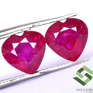 Puede incluir: Dos piedras preciosas de rubí en forma de corazón sostenidas por unas pinzas. Los rubíes son de color rosa intenso y tienen un corte facetado.